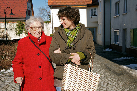 Eine ältere Frau läuft eingehakt neben einer jüngeren Frau. Die jüngere Frau trägt eine Einkaufstüte.